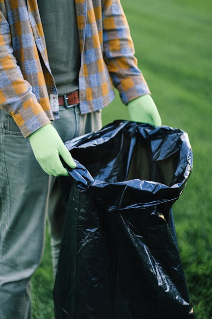 A person standing outdoors on a grassy area, wearing a yellow and grey checkered jacket, green gloves, and grey trousers with a brown belt, is holding open a large black rubbish bag lined with a yellow inner lining. The individual appears to be involved in waste collection or disposal, with the focus on their hands and the open rubbish bag, ready for debris or garden waste. The background consists of a green lawn, suggesting a garden or park setting, and the scene has natural daylight, highlighting the textures of the gloves, the bag, and the person's clothing. The image relates to waste management services, illustrating the process of independent rubbish collection or on-site clearance, potentially supporting content about private waste disposal options offered by companies like Rubbish Clearance Watford, particularly in the context of garden or household rubbish removal in Watford or similar areas.