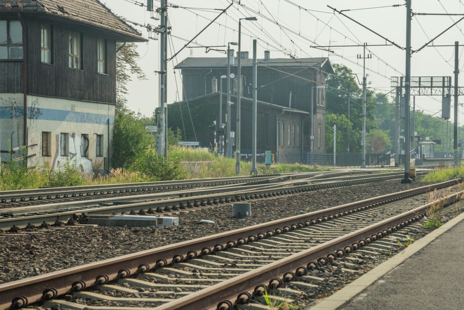 The image shows a section of railway tracks in an urban area, with multiple parallel sets of metal rails embedded in dark gravel and wooden sleepers. To the left, there is a weathered two-story building with dark wooden siding on the upper floor and a light-colored base, featuring small, irregularly shaped windows and signs of aging such as peeling paint. Overgrown grass, weeds, and low shrubs are visible along the edges of the railway and the building, indicating limited maintenance in the area. In the background, a larger dark brick or concrete structure with multiple windows and a flat roof is partially obscured by trees and railway infrastructure. Overhead, a network of electric wires and supporting poles stretches across the scene, suggesting the railway is electrified. The lighting appears natural and slightly hazy, creating a muted, industrial atmosphere. The scene is situated outdoors, likely near a station or yard, and while it relates to the broader context of railway environments, it does not depict any active rubbish clearance or collection process. The image aligns with private or alternative waste handling contexts found in urban infrastructure settings, consistent with services like those provided by Rubbish Clearance Watford for waste removal from such areas.