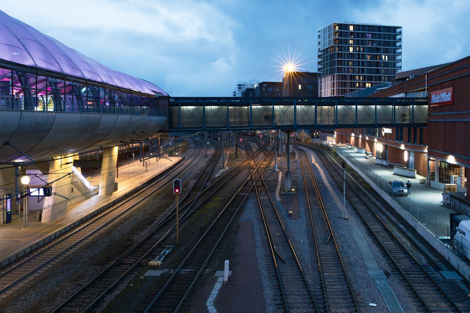 A railway yard viewed from ground level featuring multiple sets of train tracks running parallel and diverging in different directions. The tracks are made of steel rails mounted on wooden sleepers, with some sections overgrown with grass and yellow wildflowers. Overhead, there is an extensive network of electrical catenary wires supported by numerous metal poles and lattice framework, extending toward the horizon. In the background, a stationary train with white and red carriages is visible, partially obscured by the track infrastructure. The sky above is partly cloudy with patches of blue, casting diffuse daylight over the scene. On the right edge, a concrete or brick structure with faint weathering is partially visible. This scene is typical of a busy rail freight or passenger yard, reflecting an environment where rail operators like Rubbish Clearance Watford may encounter old equipment or infrastructure during site inspections or on-site clearance activities, emphasizing the importance of managing waste associated with transport and railway upgrades.