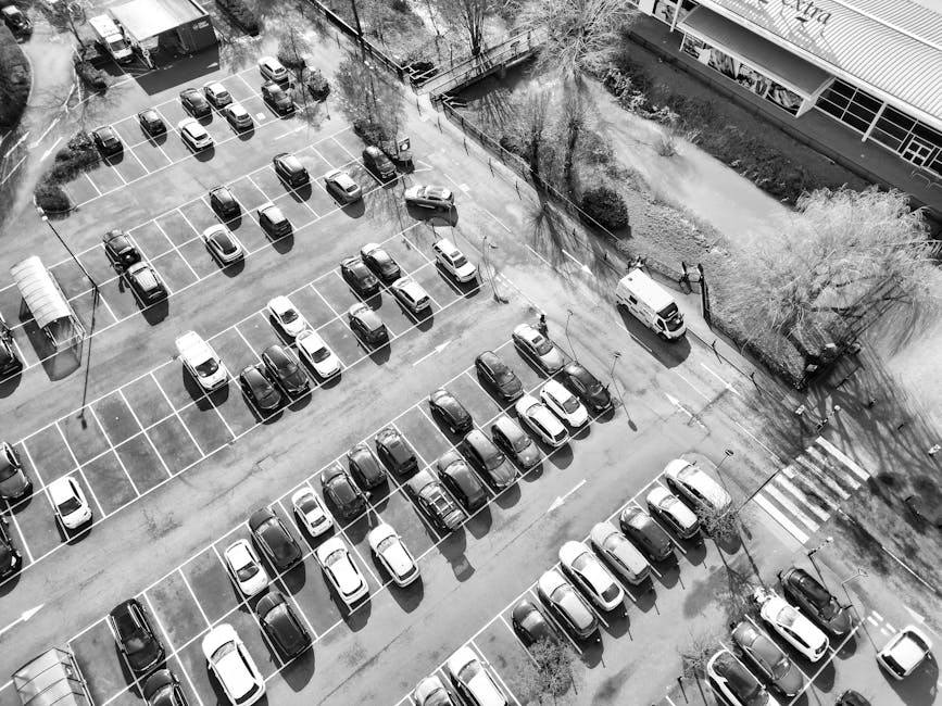 An aerial view of a large parking lot filled with rows of parked cars of various makes and models, mostly in dark and light tones, organized within marked parking bays with white lines. The parking lot is surrounded by trees casting shadows across the asphalt and adjacent to a modern building with large windows and a pavement walkway, visible at the top and right edges of the image. There is a white van parked along the curb near the trees, and a few pedestrians are walking or standing in the vicinity. The scene appears to be during daylight hours with natural light creating shadows from the trees and vehicles, and the overall environment suggests a commercial or office area where space management and parking are essential for daily operations. Though not directly related to rubbish removal services, the image showcases a typical urban or business context where waste management and on-site clearance may be relevant to maintaining the area, aligning with independent waste disposal or rubbish collection activities.