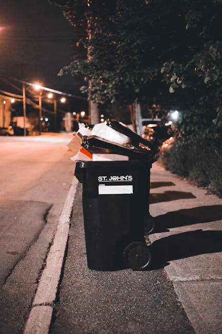 An open black wheeled waste bin positioned on the edge of a street at night, with its lid raised and overflowing with a mixture of cardboard boxes, paper, and various flattened packaging materials. The bin features a white label with the inscription 'ST. JOHN'S' and is situated on a paved sidewalk adjacent to a curb. In the background, faint streetlights illuminate the deserted road, with trees and shrubbery casting shadows on the pavement. The scene suggests an informal or alternative rubbish disposal setting, possibly associated with private waste management, in alignment with services offered by Rubbish Clearance Watford for rubbish removal and on-site clearance tasks. The surrounding environment includes street lighting, a few parked vehicles in the distance, and the natural texture of the foliage on the right side of the image, providing a neutral, professional context for waste collection and disposal activities outside conventional public collection points.