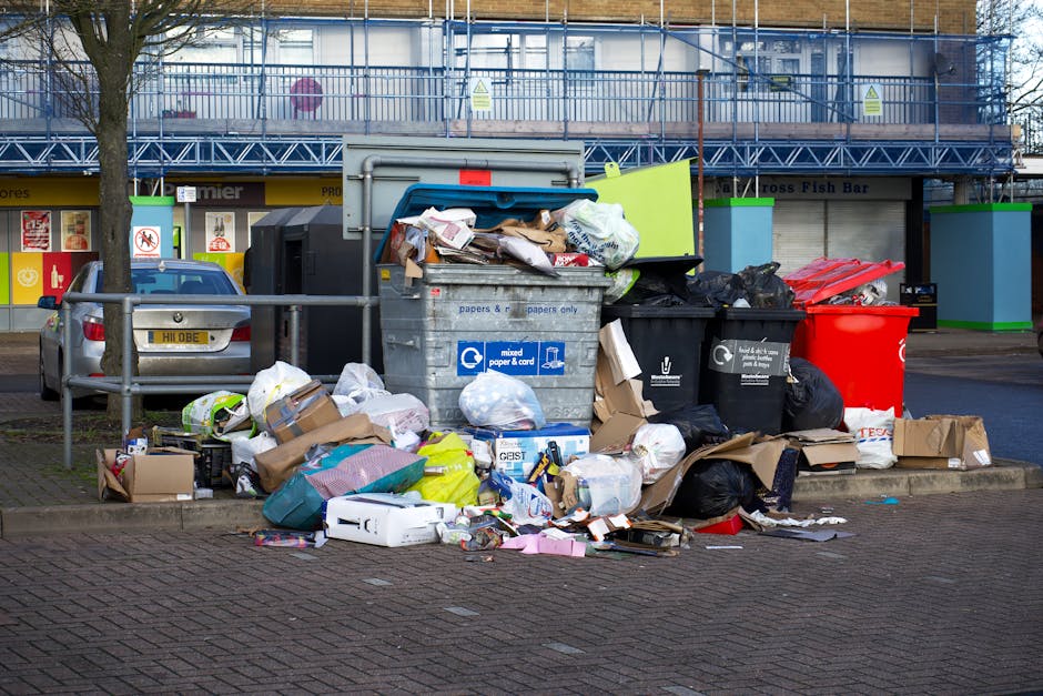 A large collection of mixed waste and rubbish bags, including black, black, and red plastic rubbish bags, cardboard boxes, and loose paper, are piled outside a council-approved recycling bin designated for mixed paper and cardboard. The waste is situated on a paved area with a concrete curb in front, adjacent to a parking space with a silver hatchback bearing a yellow license plate. Behind the waste, there is a building with a storefront, partially obscured by a metal railing, and a tree trunk is visible on the left side of the image. The recycling bin appears to be overfilled, with items spilling onto the ground, and is located in an urban environment that includes a small retail shop or fast-food outlet with window signage, indicating a commercial area. The scene reflects an instance of improper waste disposal, which could be addressed by private rubbish removal services such as those offered by Rubbish Clearance Watford, to manage estate clearances and local rubbish collection needs efficiently.