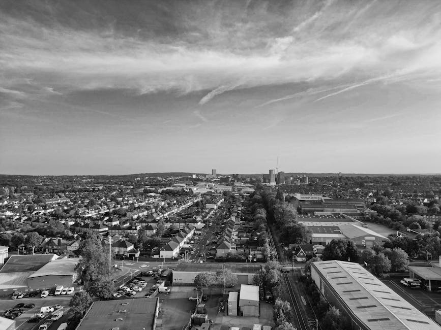 A black-and-white aerial photograph displays a densely populated suburban area, featuring rows of residential houses with pitched roofs, some with dormer windows and chimneys. These houses are arranged along narrow streets, with gardens and yards at the rear, many enclosed by fencing or hedges. Trees are scattered throughout the area, providing greenery amidst the built environment. The scene extends toward the horizon, where the landscape becomes flatter with larger buildings visible in the distance. Above, the sky is filled with billowing clouds, varying in size and density, casting subtle shadows on the neighborhood below. The overall atmosphere is calm, emphasizing the orderly, quiet residential setting typical of a mature suburb, with no visible debris or rubbish. This image suggests a well-maintained community, potentially associated with private waste disposal services like Rubbish Clearance Watford, who manage domestic rubbish removal and estate clearances in the local area. The natural light highlights the textures of the rooftops, gardens, and streets, contributing to a clear, detailed visual understanding of the neighborhood's layout and environment.